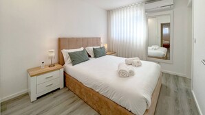 Une chambre à coucher moderne avec une tête de lit en bois, un grand lit recouvert de draps blancs, une table de chevet avec une lampe et un miroir accroché au mur.