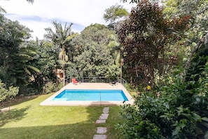 Una piscina rectangular con un camino de piedra que la conduce, rodeada de abundante vegetación y árboles.