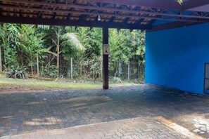 An open covered area with a brick floor, surrounded by greenery and a blue wall. - Casa ampla com piscina e churrasqueira  (Carapicuíba)