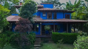 Una casa de dos pisos con techo de tejas, rodeada de abundante vegetación y un pequeño jardín.