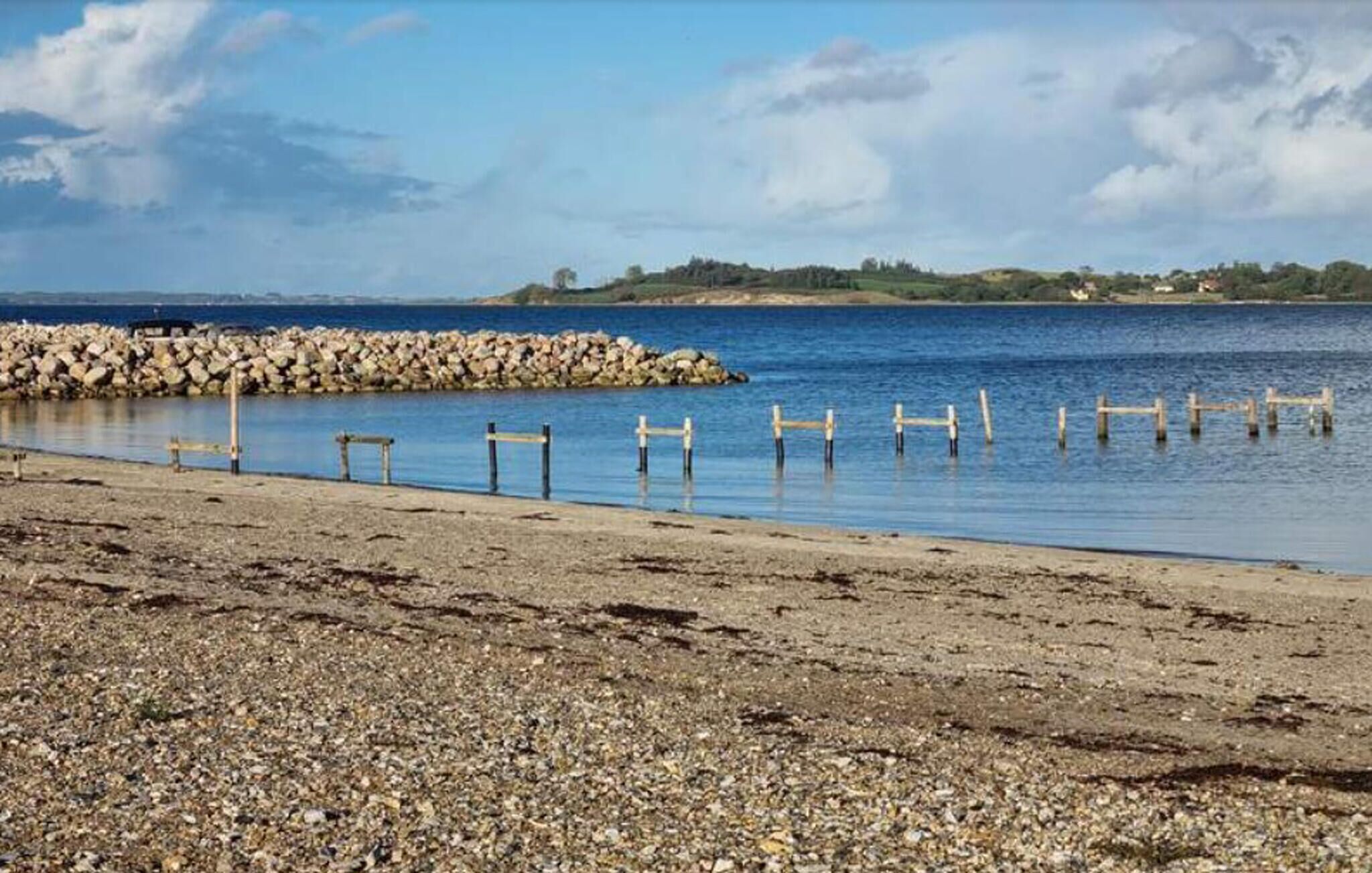 A pebbly beach with wooden posts in the water, a rocky breakwater, and a distant shoreline with greenery.