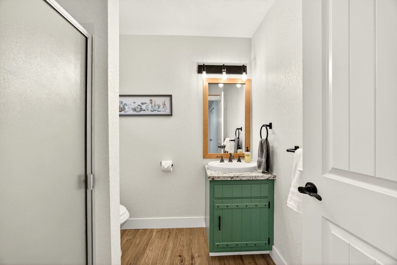 A bathroom with a green vanity, a white toilet, a framed artwork on the wall, and a mirror with overhead lighting.