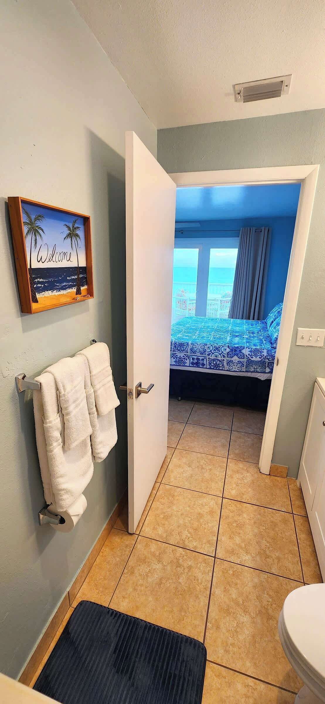 Ein Badezimmer mit Toilette, einem Handtuchhalter mit Handtüchern, einem begrüßungsschild im Bilderrahmen und Blick auf ein Schlafzimmer mit Bett und Vorhängen.