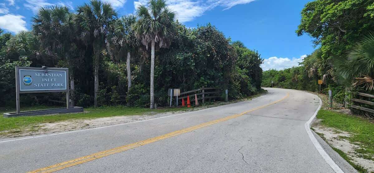 Una carretera que lleva al Parque Estatal Sebastian Inlet, con un cartel de señalización, rodeada de abundante vegetación y palmeras.