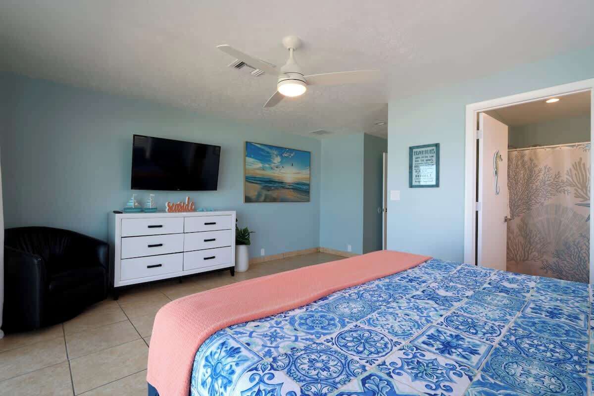 Une chambre à coucher comprenant un lit, une télévision, un ventilateur de plafond, une commode et une porte donnant sur une salle de bain.