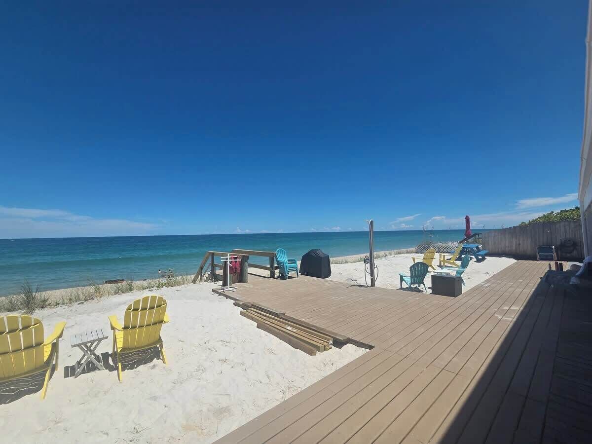 Eine Strandterrasse mit gelben Adirondack-Stühlen, einem überdachten Grill und einem Holzdeck, das bis zum Sand reicht.