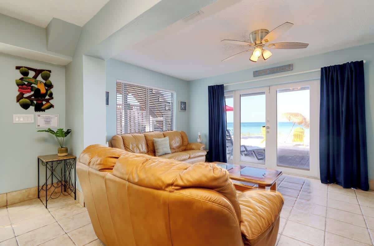A living room with a leather sofa, a ceiling fan, a small table, and a view of the beach through large windows.