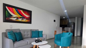 Ein modernes Wohnzimmer mit einem grauen Sofa, türkisfarbenen Kissen und zwei weißen runden Tischen. An der Wand hängt ein farbenfrohes abstraktes Gemälde.