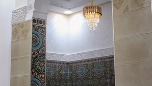 Une pièce avec un mur recouvert de carreaux décoratifs, un lustre suspendu et un espace salon aménagé en alcôve.