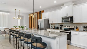 Una cocina moderna con armarios blancos, electrodomésticos de acero inoxidable, una isla de granito con taburetes y luminarias colgantes.