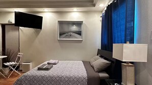 A modern hotel room with a bed, a desk with a lamp, a flat-screen TV mounted on the wall, and a window with blue curtains. - Isla Verde Beach Studio with Pool & Parking! A2 (Carolina)