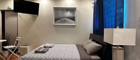 A modern hotel room with a bed, a desk with a lamp, a flat-screen TV mounted on the wall, and a window with blue curtains.