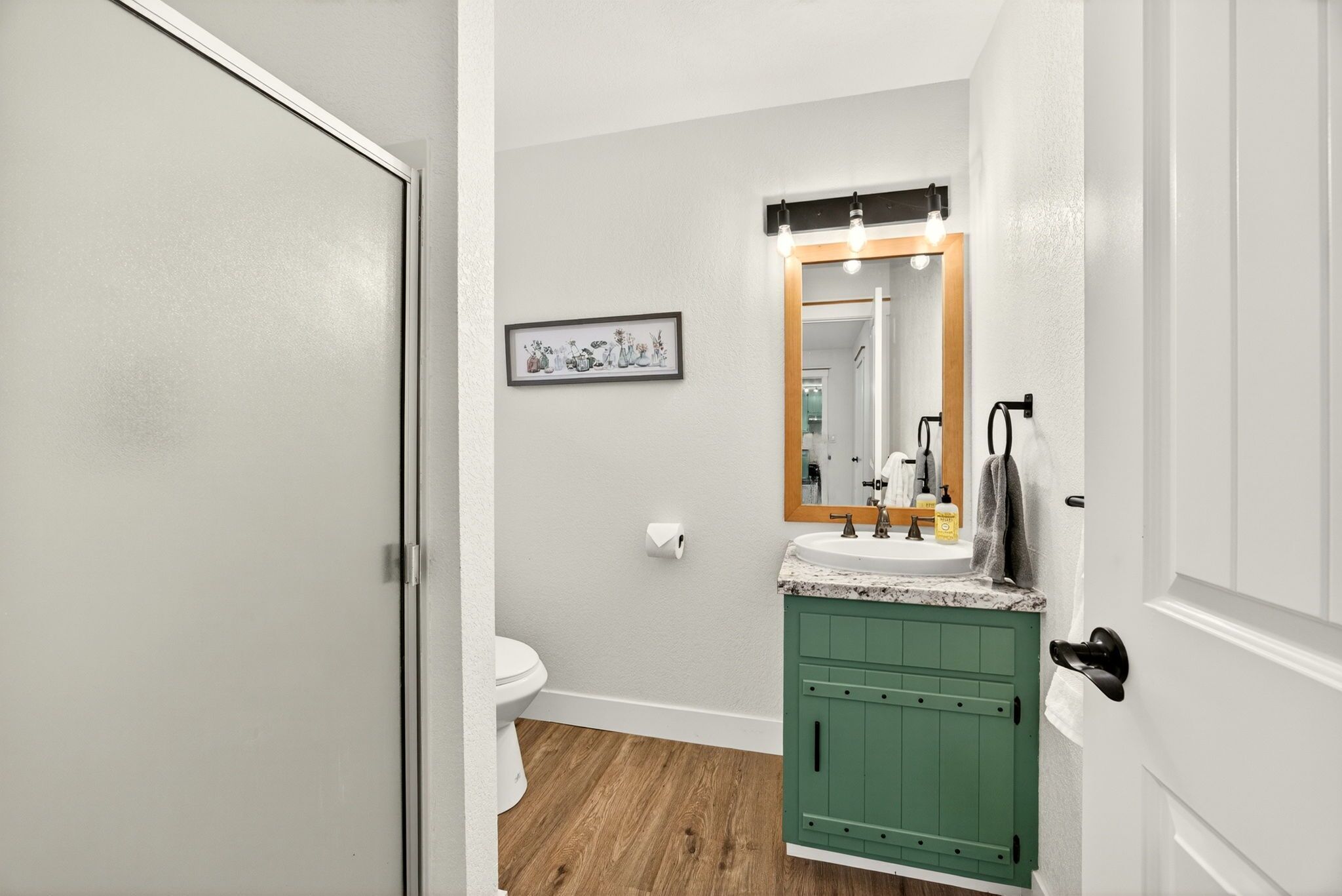 A bathroom with a toilet, green cabinets, a sink with a mirror, and a framed picture on the wall.