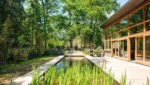 Ein modernes Gebäude mit großen Glasfenstern, einer Holzterrasse und einem Teich mit Wasserpflanzen.