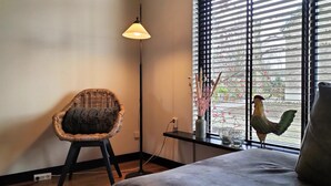 Una habitación con una silla de mimbre, una mesa con un jarrón y una decoración de gallo cerca de una ventana con persianas.
