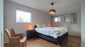 Un dormitorio con cama, una silla, una mesita de noche con lámpara, una lámpara colgante y una ventana con vista a edificios.
