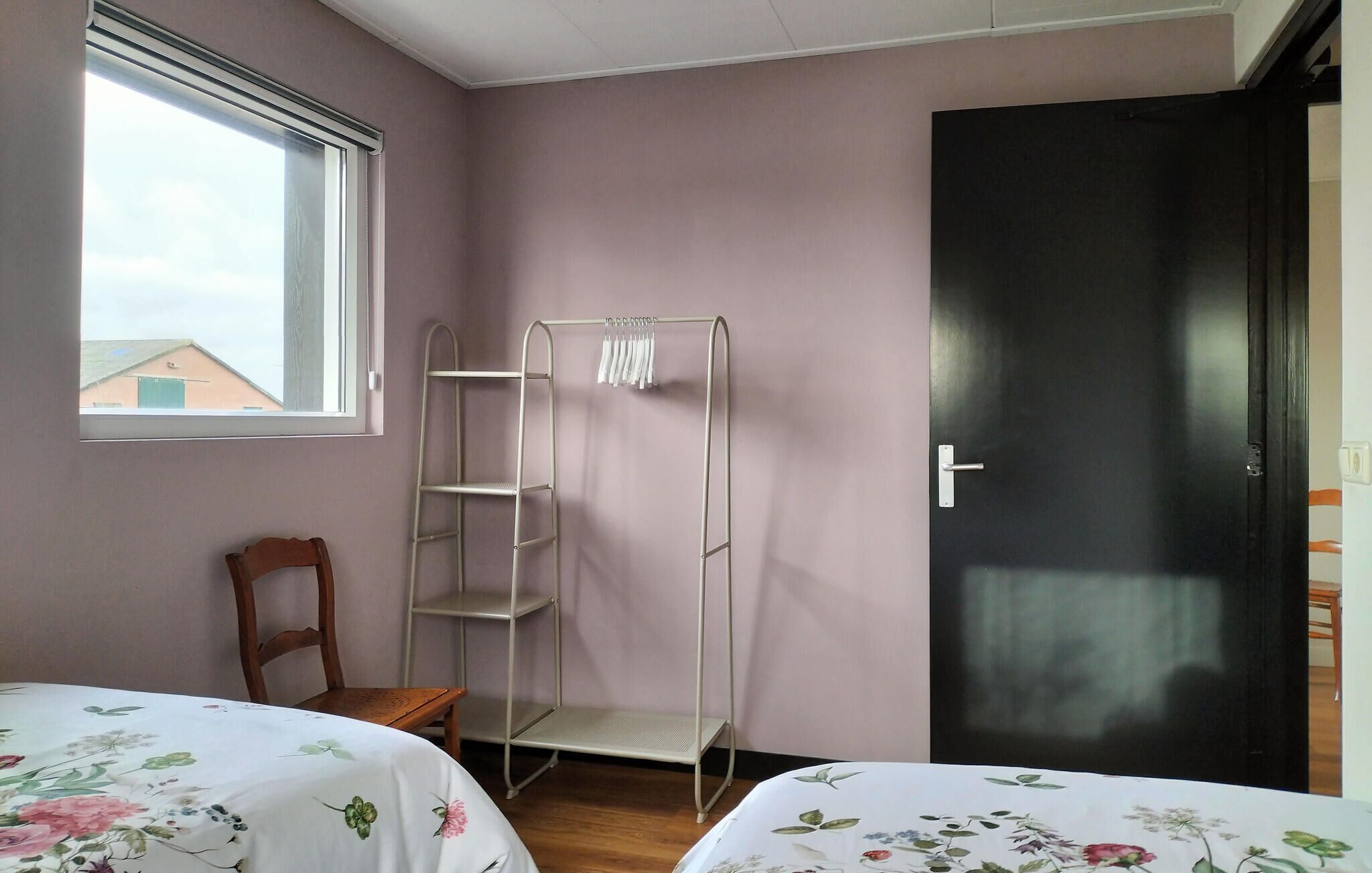 Ein Schlafzimmer mit zwei Betten, einem Metallregal, einem Holzstuhl und einem Fenster mit Blick auf ein Gebäude.