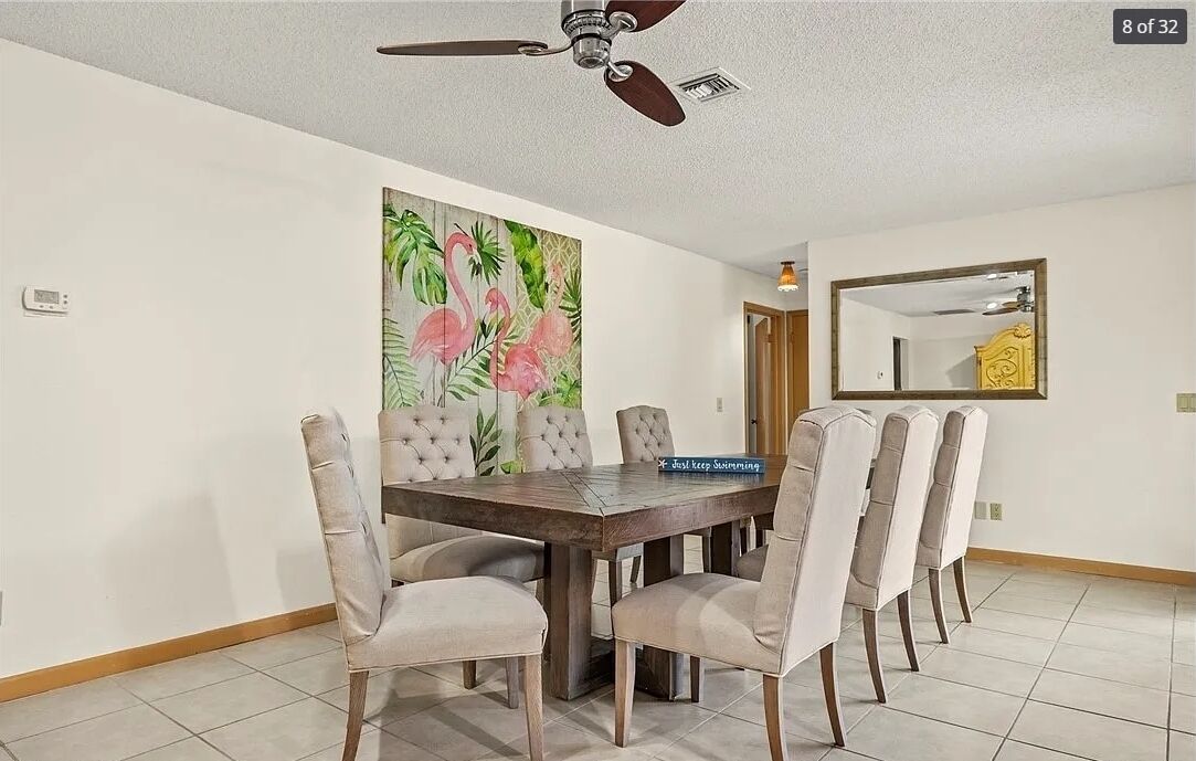 Comedor con mesa de madera y seis sillas, ventilador de techo, un cuadro grande de flamencos y un espejo en la pared.