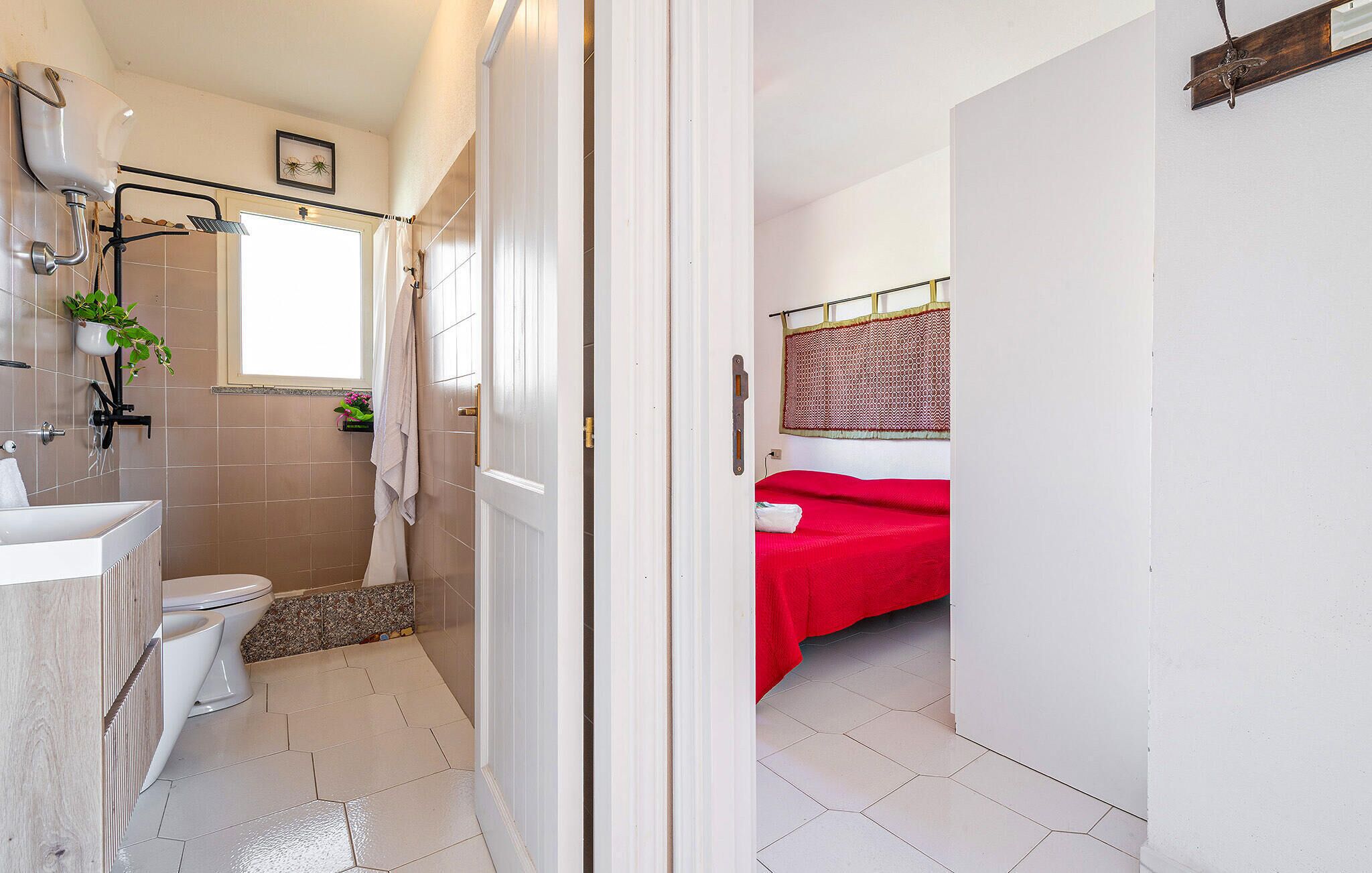 A bathroom with a white sink, toilet, and shower. There is a red bed in the adjacent room.