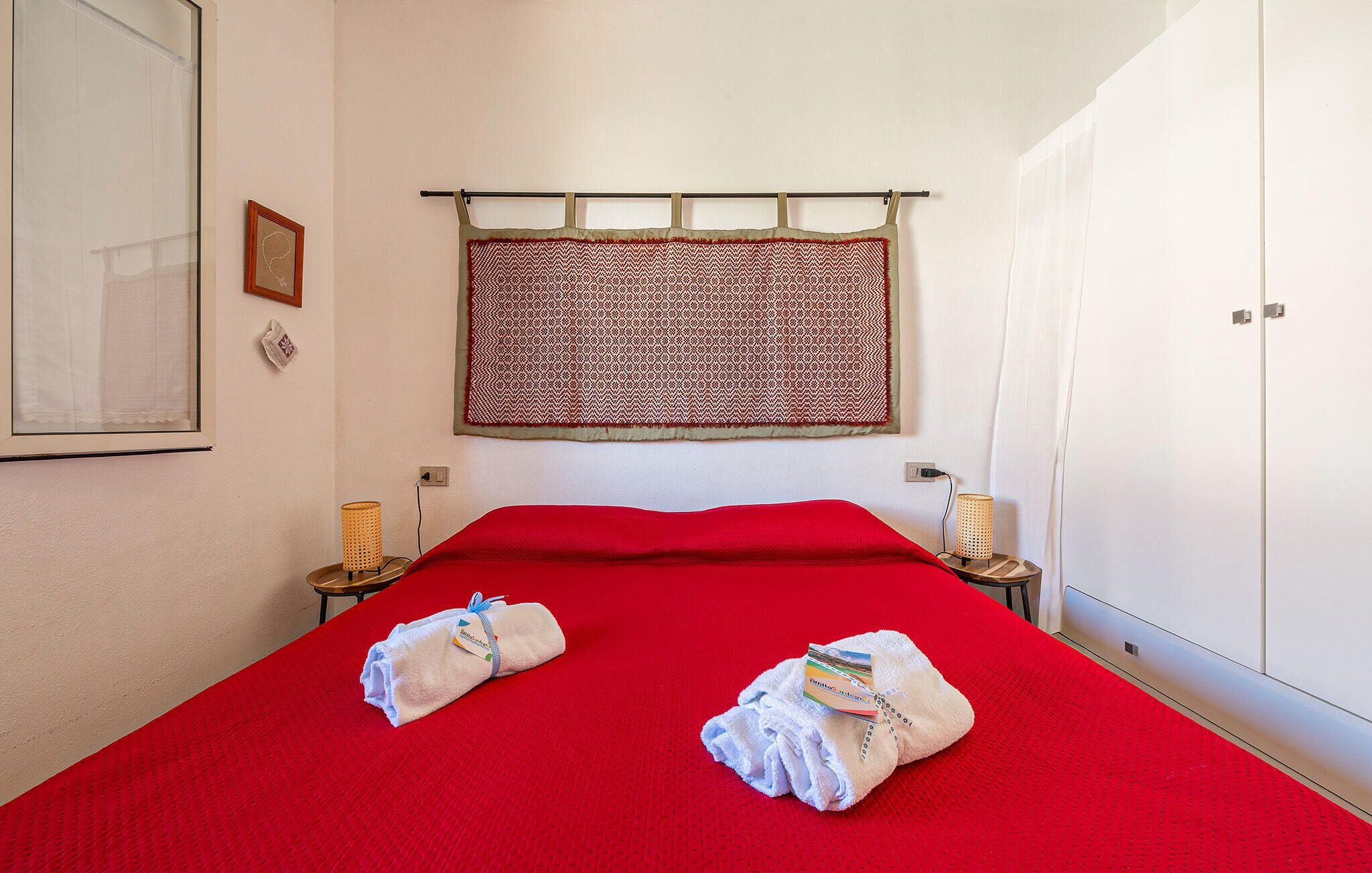 A hotel room with a red bedspread, two towels on the bed, a patterned curtain, and a small table with a lamp.