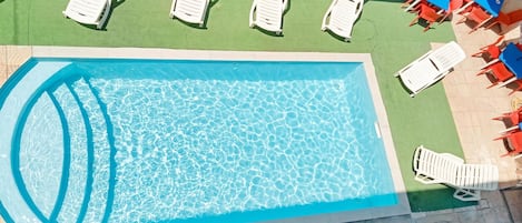 Suorakulmainen uima-allas, jossa on kirkasta, sinistä vettä, ja sen ympärillä on laatoitettu allasterassi. Terrassilla on useita valkoisia aurinkotuoleja sekä muutama punainen ja sininen tuoli.