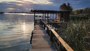 Un muelle de madera que conduce a un área de descanso techada con vista al agua y al cielo.