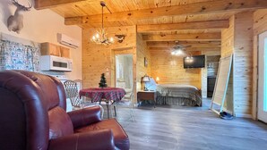 Interior acogedor de una cabaña de madera con un sillón de cuero, una mesa de comedor con mantel a cuadros, un pequeño árbol de Navidad, una cama con manta a rayas, una cabeza de ciervo montada y un televisor montado en la pared.