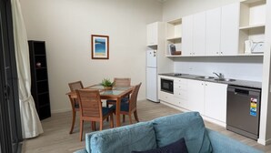 Een compacte keuken met witte kasten, een inbouwoven en een vaatwasser. Er is een eettafel met stoelen en een kleine potplant.