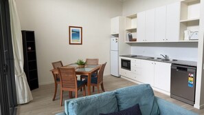 A compact kitchen with white cabinets, a built-in oven, and a dishwasher. There is a dining table with chairs and a small potted plant. - Baden 79 - Rainbow Shores (Rainbow Beach)