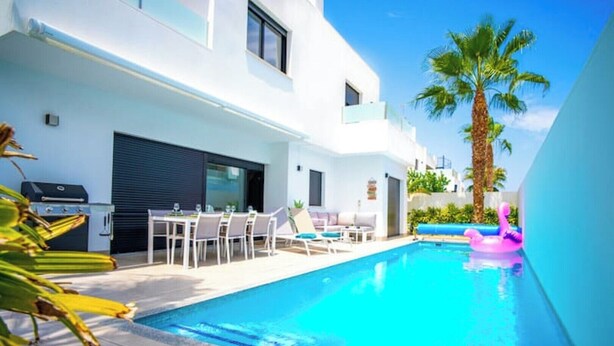 Un área de piscina moderna con una piscina azul cristalina, una mesa de comedor con sillas y una tumbona.
