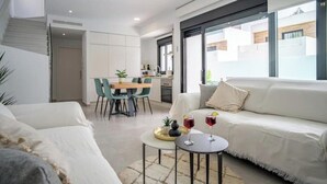 Una sala de estar moderna con un sofá blanco, un comedor con una mesa y sillas de madera, y una cocina al fondo.