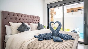 Un dormitorio moderno con un cabecero capitonado, una cama grande con colcha color beige y un arreglo de toallas en forma de corazón sobre la cama.
