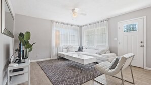 Ein modernes Wohnzimmer mit einem weißen Sofa, einem Couchtisch und einem weißen Sessel. Es gibt einen Deckenventilator, eine Pflanze und ein Fenster mit Vorhängen.