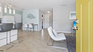 Eine moderne Küche mit weißen Schränken, ein Essbereich mit weißem Tisch und Stühlen sowie ein Wohnbereich mit einer weißen Couch und einem Wanddekor.