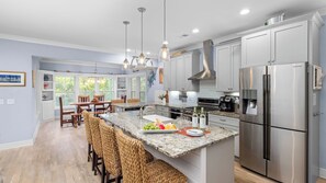 A modern kitchen with a stainless steel refrigerator, granite countertops, and a dining area with wicker chairs. - Folly Beach Getaway Near Sand Covered Lounge, BBQ & Outdoor TV Tidecrest by Avantstay (Folly Beach)
