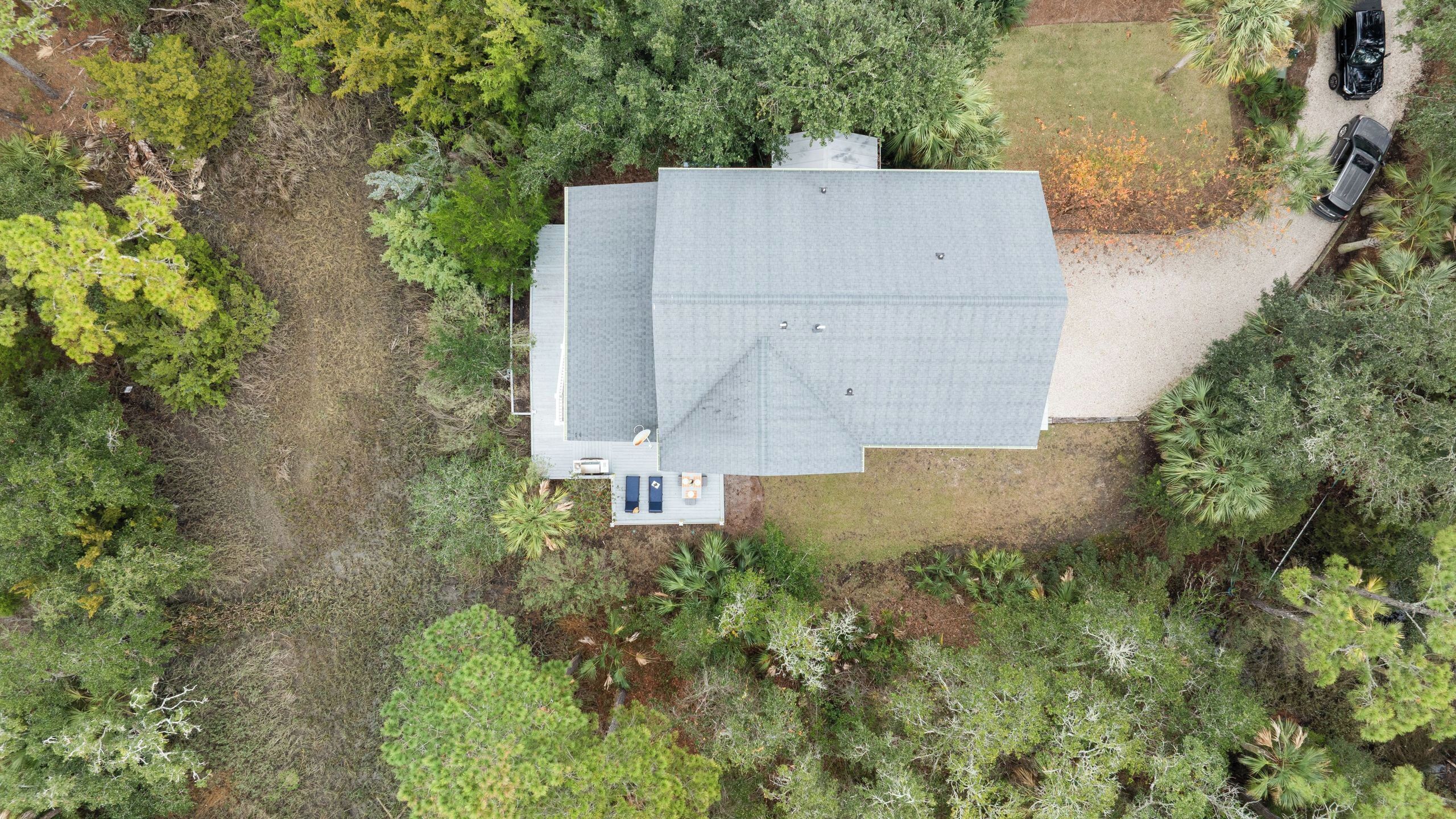 Vista aérea de uma casa com um telhado grande, cercada por vegetação densa e uma entrada com dois carros.