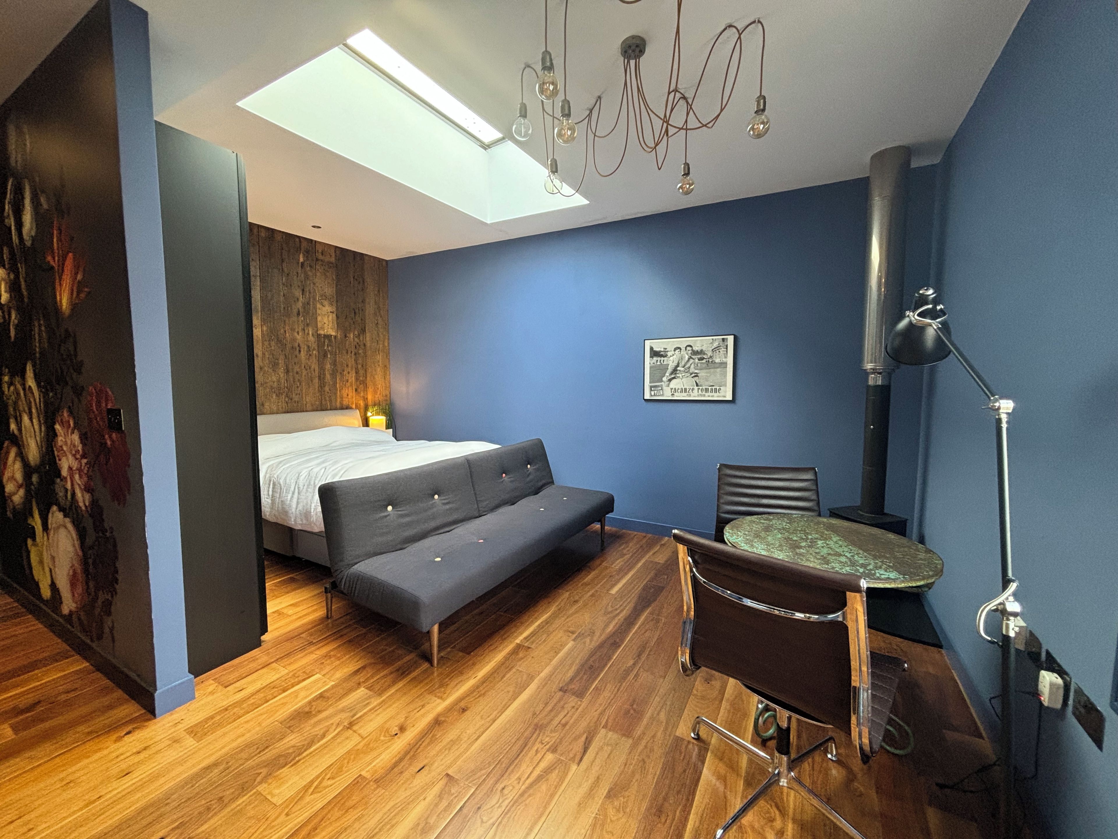 A room with a bed, a sofa, a desk, and a chair. There is a ceiling light and a wall-mounted lamp.