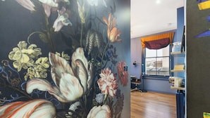 Ein Zimmer mit einer großen floralen Wandmalerei, einem Fenster mit orangefarbenen Vorhängen sowie einem Bücherregal mit Büchern und einer Lampe.