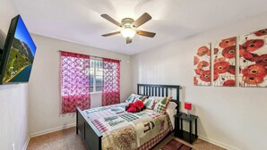 Ein Schlafzimmer mit einem Bett, einem Deckenventilator, einem Fenster mit floralem Vorhang, einem Bild an der Wand und einem kleinen Nachttisch mit einer Lampe.