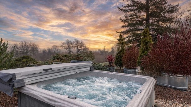 Un jacuzzi con revestimiento de piedra, rodeado de plantas en macetas y árboles, con el cielo de un atardecer de fondo.