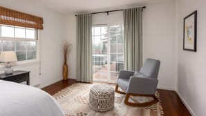 Un dormitorio con cama, una silla, mesita de noche con lámpara, ventana con cortinas y vistas al exterior.