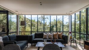 A modern living room with a large window, a sofa, a coffee table, and a lamp. - The Palm Beach Treehouse (Waiheke Island)