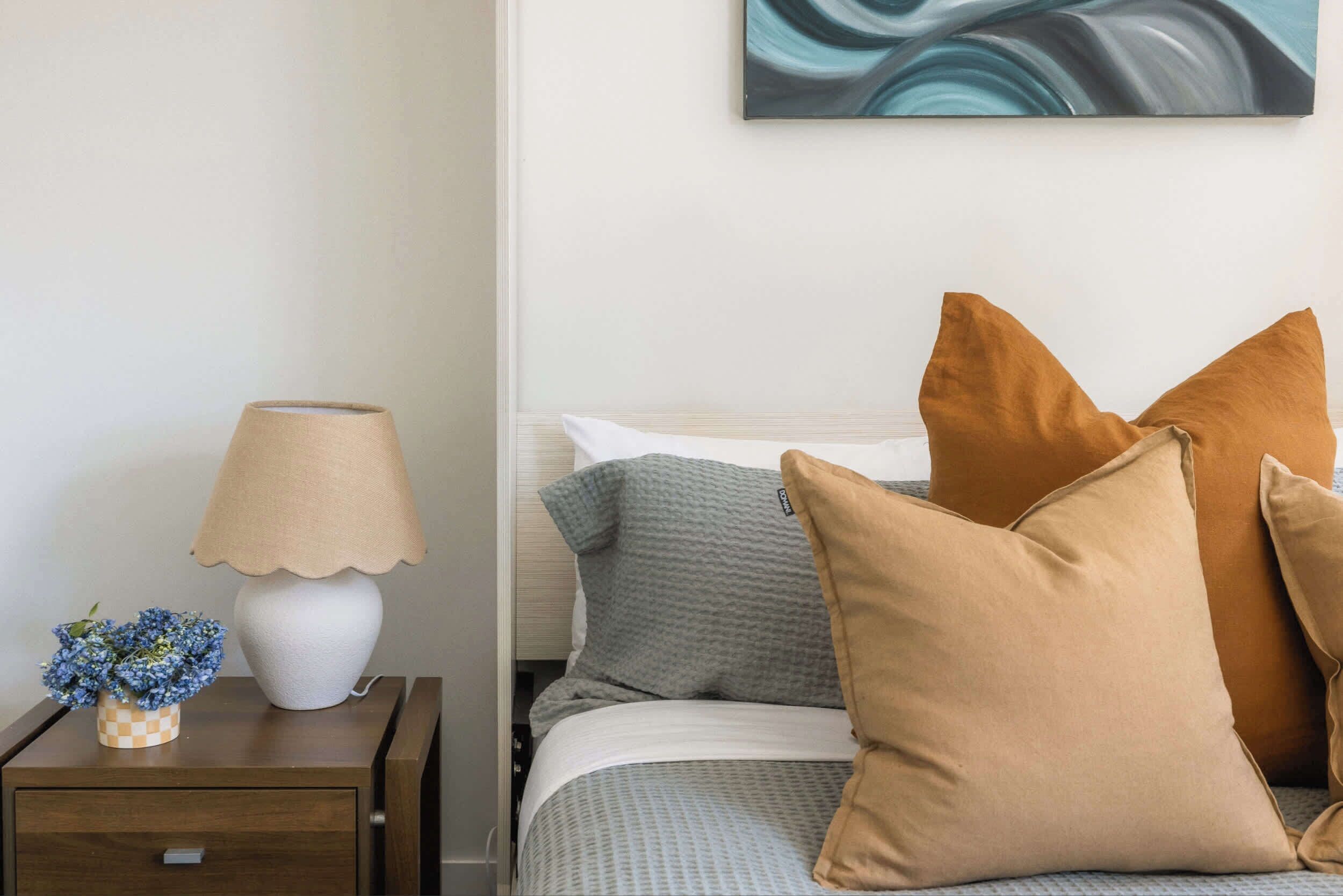 Een netjes opgemaakt bed met meerdere kussens, een bedlampje en een klein decoratief vaasje met bloemen op een houten nachtkastje.