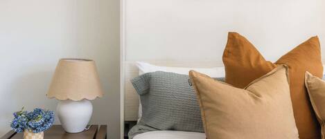 Een netjes opgemaakt bed met meerdere kussens, een bedlampje en een klein decoratief vaasje met bloemen op een houten nachtkastje.