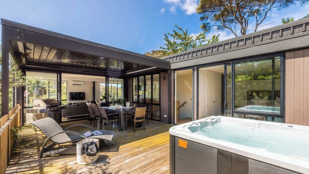 A modern outdoor deck with a hot tub, seating area, and a covered patio. - The Palm Beach Treehouse (Waiheke Island)