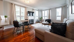 Una sala de estar moderna con un sofá, una mesa de comedor con sillas, un televisor de pantalla plana y ventanas grandes con cortinas.