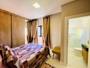 Ein Schlafzimmer mit Bett, Vorhängen, einem kleinen Hocker und einem durch eine offene Tür einsehbaren Badezimmer.