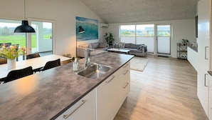 Cozinha moderna com uma ilha grande, área de jantar com mesa e cadeiras, sala de estar com sofá e janelas amplas com vista para o exterior.