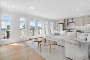 Un salon spacieux avec un grand canapé blanc, une table basse en bois et une vue sur l’océan grâce à de grandes fenêtres.