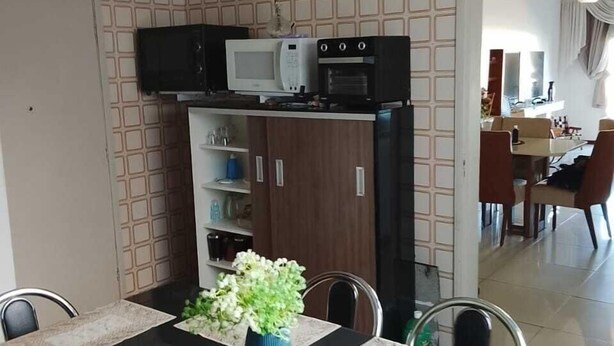 A dining area with a black table, white placemats, and a blue vase with flowers. There is a microwave and oven above the counter. - apartamento de temporada  (Uruguaiana)